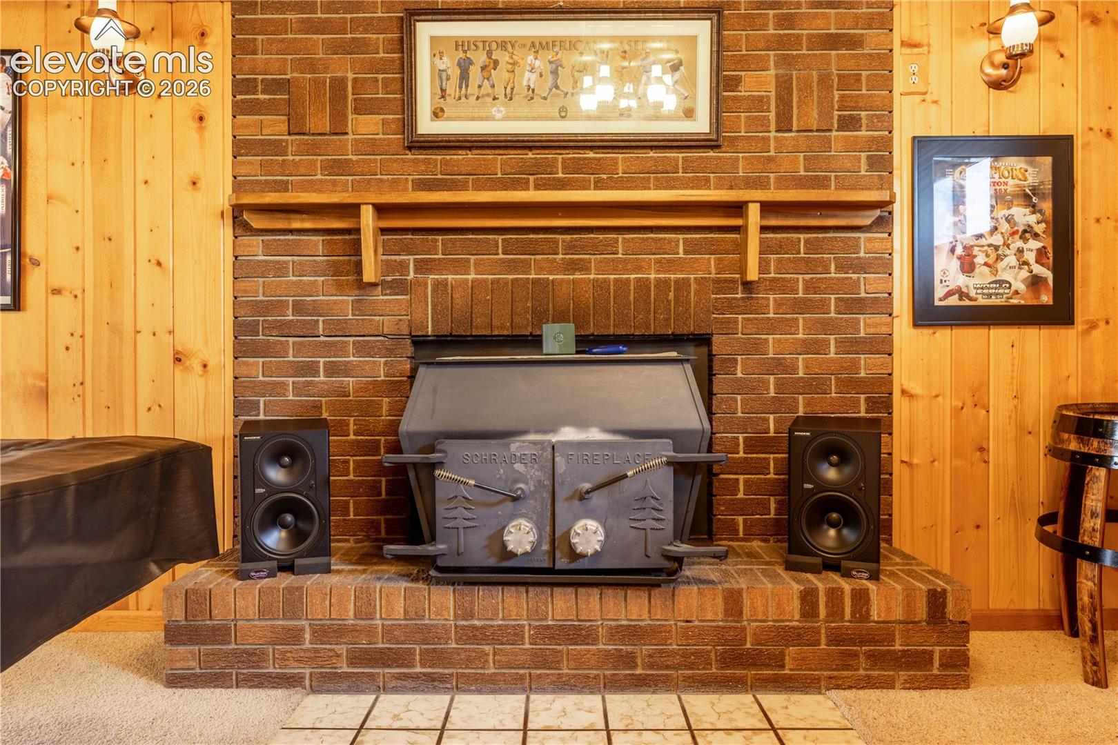 Floor-to-ceiling brick fireplace surround featuring a built-in metal wood stove, wood-finished wall paneling, a wood mantel, and dual wall-mounted light fixtures