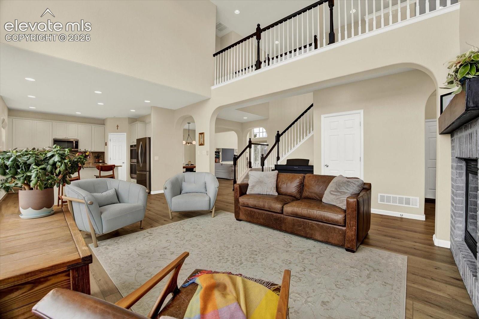 Open-concept floor plan featuring soaring double-height ceilings, a floor-to-ceiling masonry fireplace, recessed lighting, wood-finish flooring, and a decorative staircase with a spindle railing
