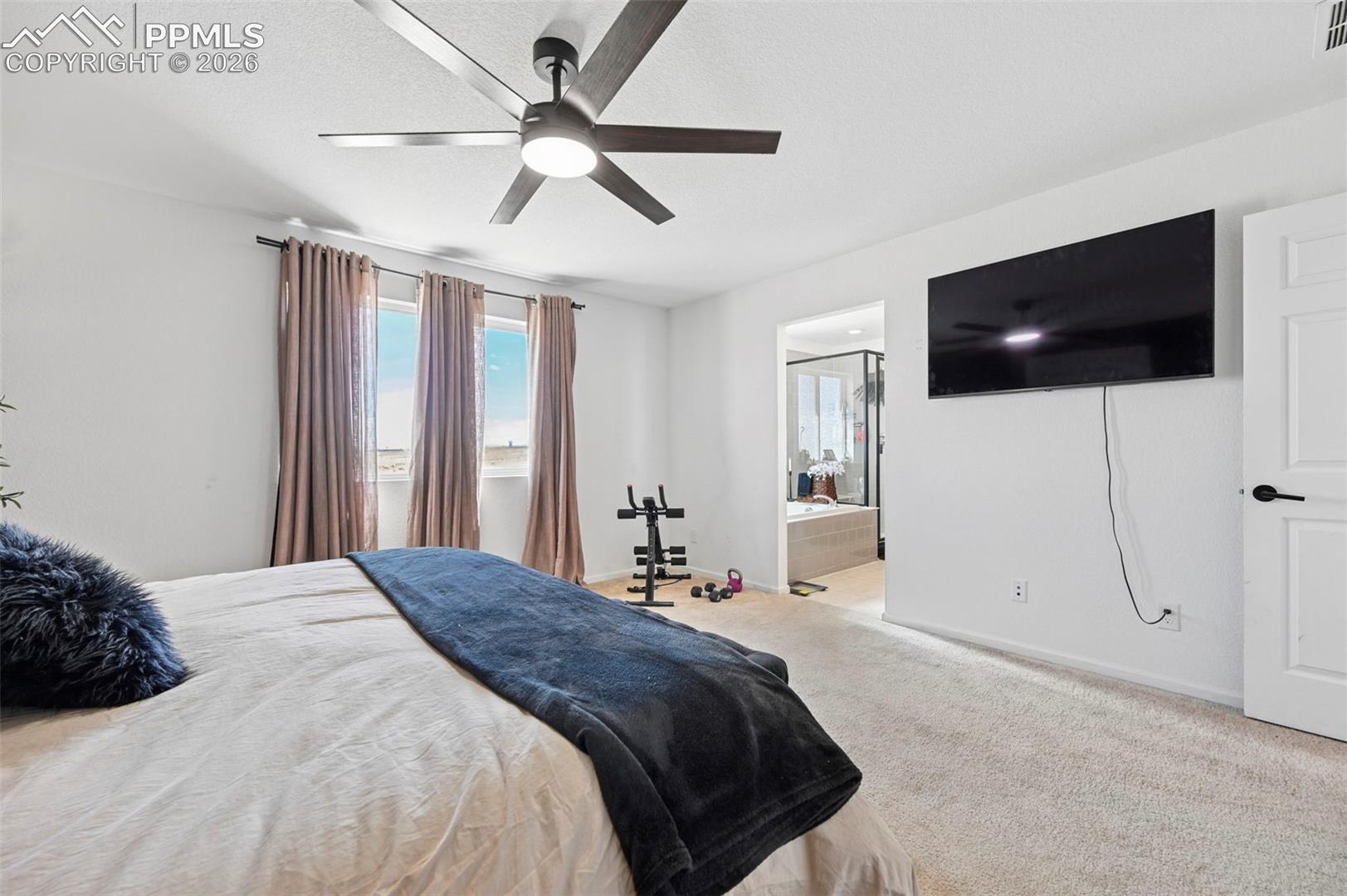 Carpeted bedroom featuring a contemporary ceiling fan with lighting, three windows draped with neutral curtains, and an entryway to an adjoining bathroom with a soaking tub and glass-enclosed shower