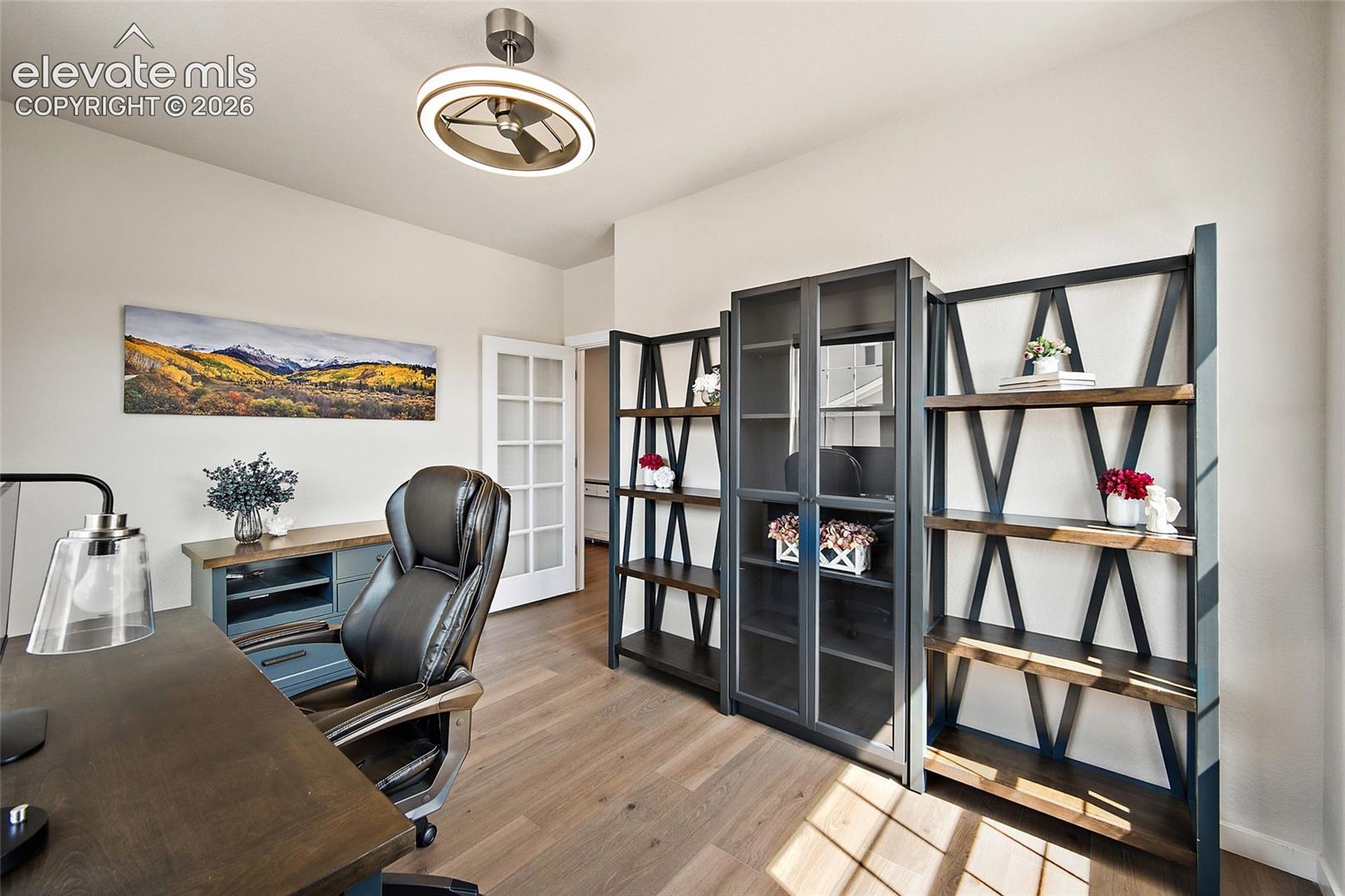 Wood-finish flooring, white paneled French doors, built-in shelving, glass-front cabinetry, and a ceiling fan with integrated lighting