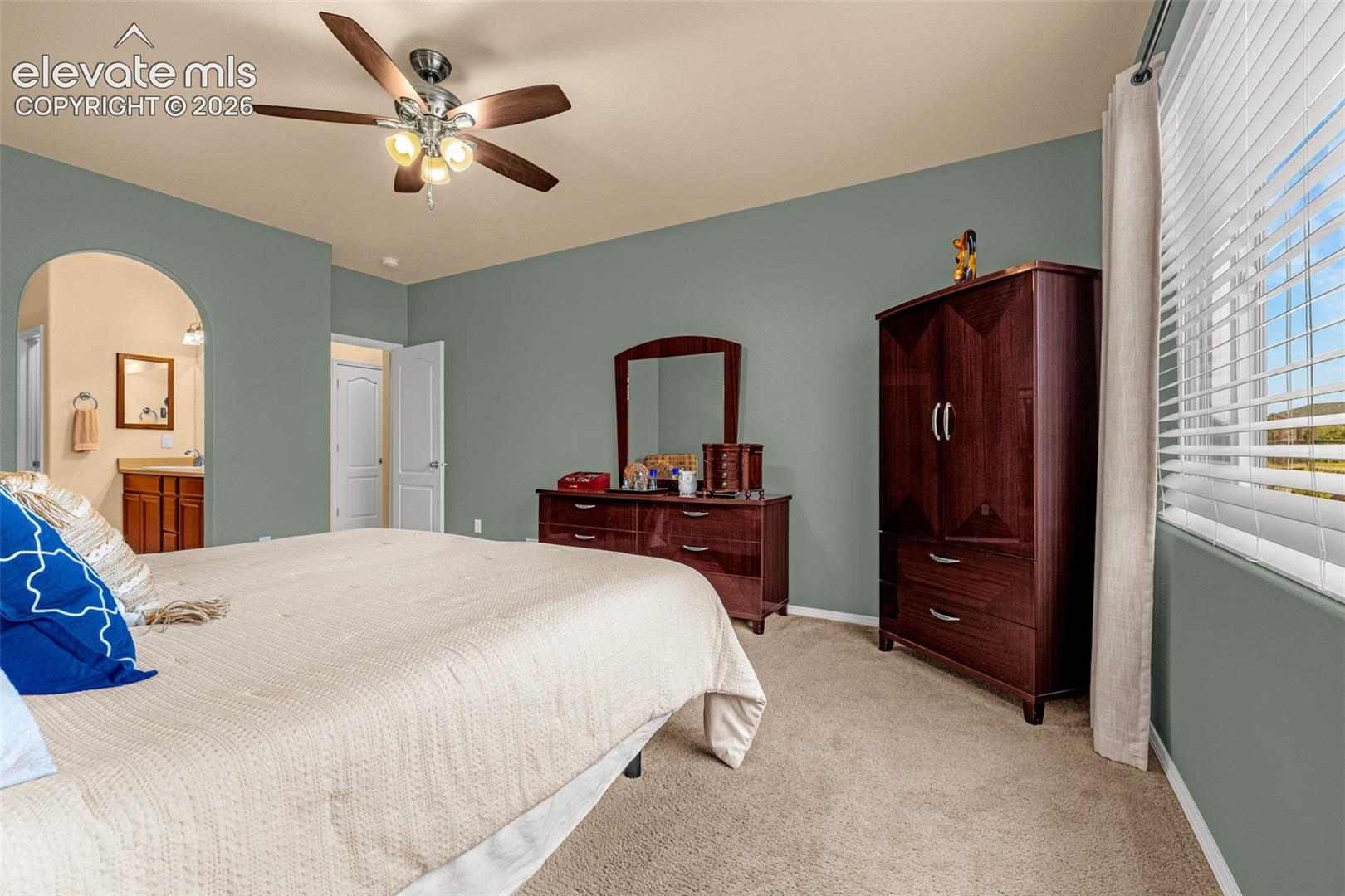 Spacious room featuring neutral carpeting, a central ceiling fan with integrated light fixture, a wide window with horizontal blinds, and a distinct arched entryway