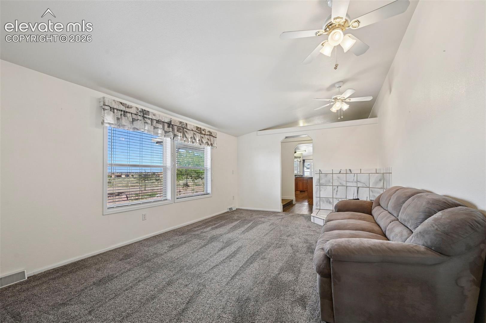 Vaulted ceilings featuring dual ceiling fans, wall-to-wall carpeting, and a fireplace surround with stone-finish tile