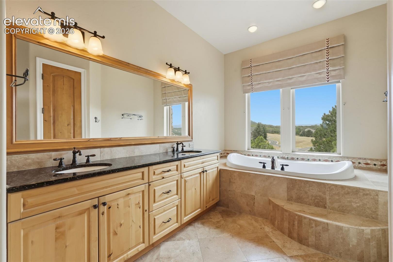 Bathroom featuring a built-in tub with tile surround, dual vanity with dark countertops, wood-finish cabinetry, and a large wall-mounted mirror