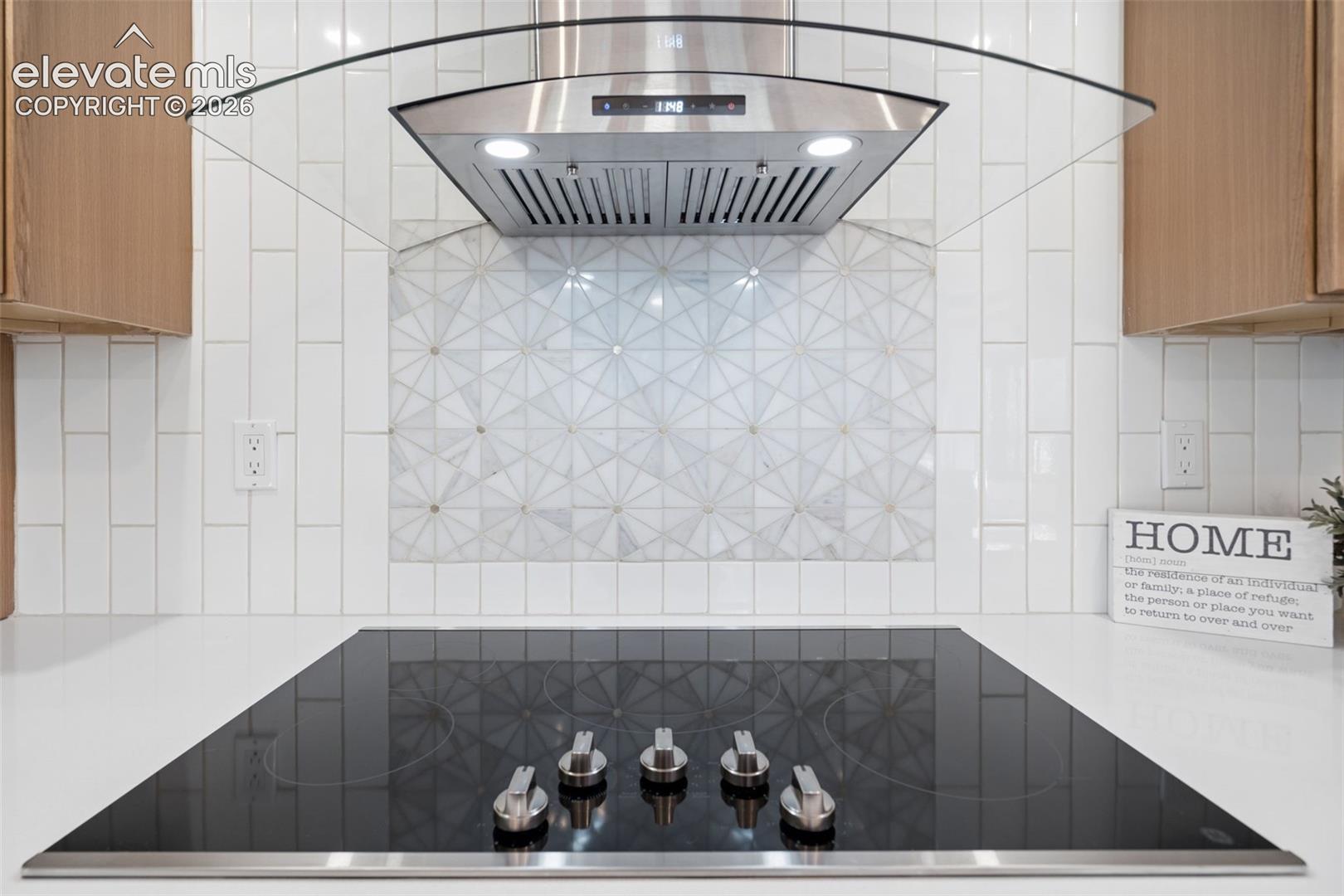 Integrated cooktop with stainless steel controls, white countertop, white vertical subway tile backsplash, decorative geometric tile inset, and stainless steel range hood with glass canopy