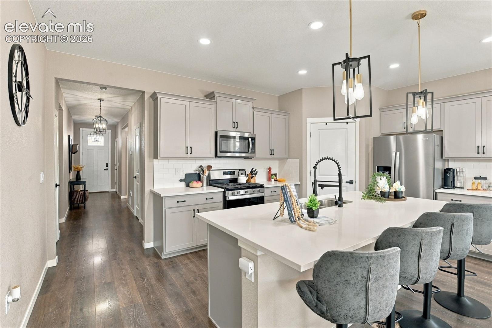 Kitchen featuring light grey shaker-style cabinetry, quartz countertops, a central island with a high-arc faucet, stainless steel appliances, and wood-finish flooring