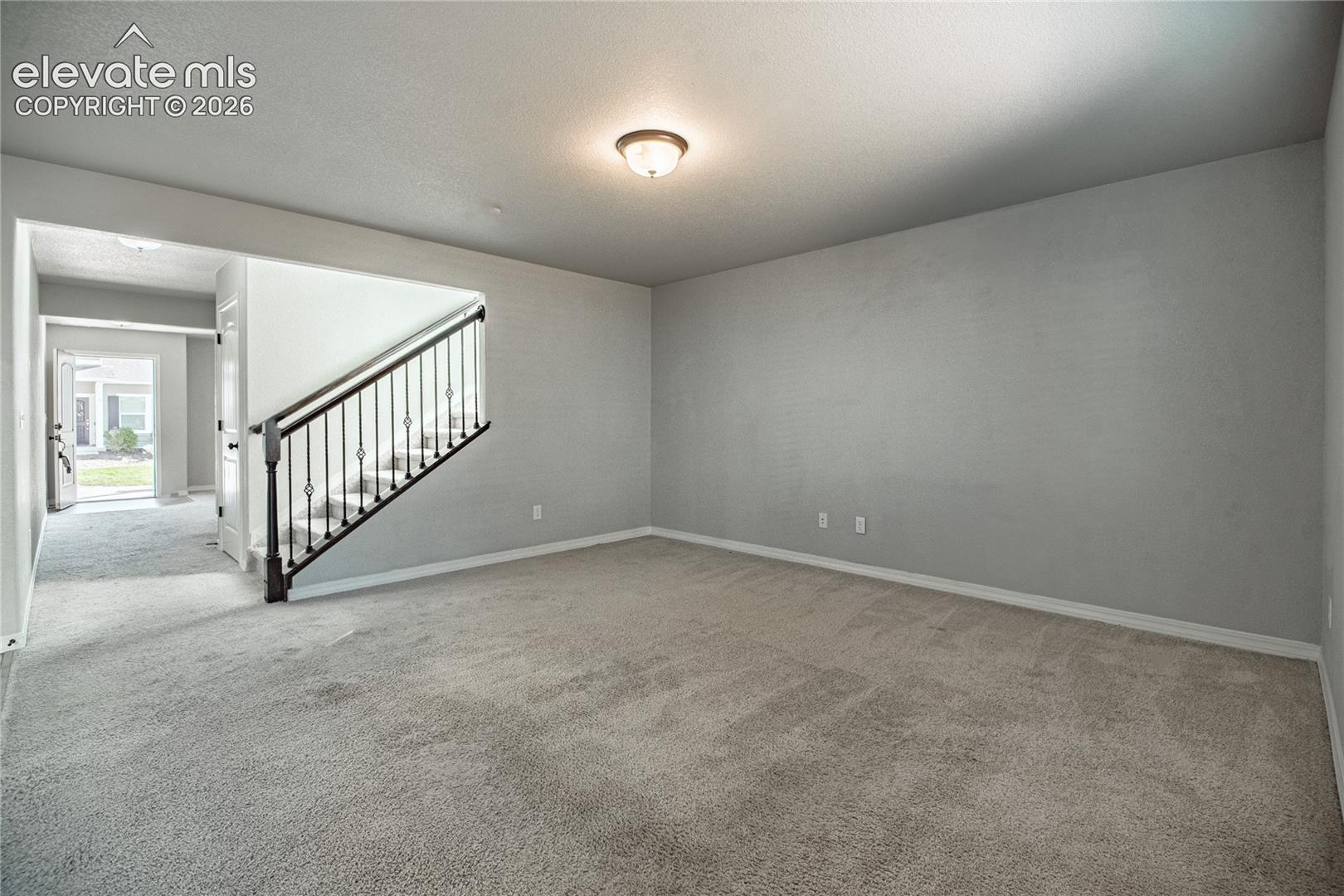 Carpeted room with light gray walls, an overhead flush-mount light fixture, and a staircase featuring a dark wood handrail and decorative metal balusters