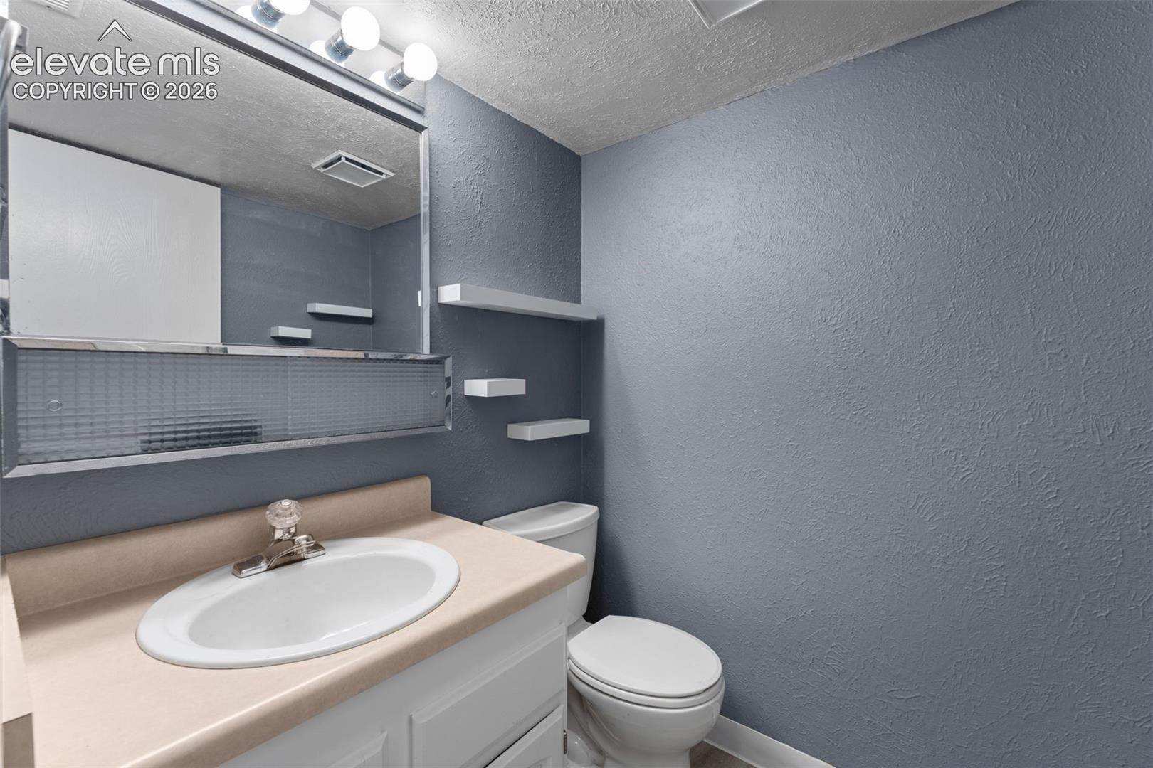 White vanity with an integrated oval sink, three wall-mounted shelves, a mirror with integrated lighting, and a white toilet