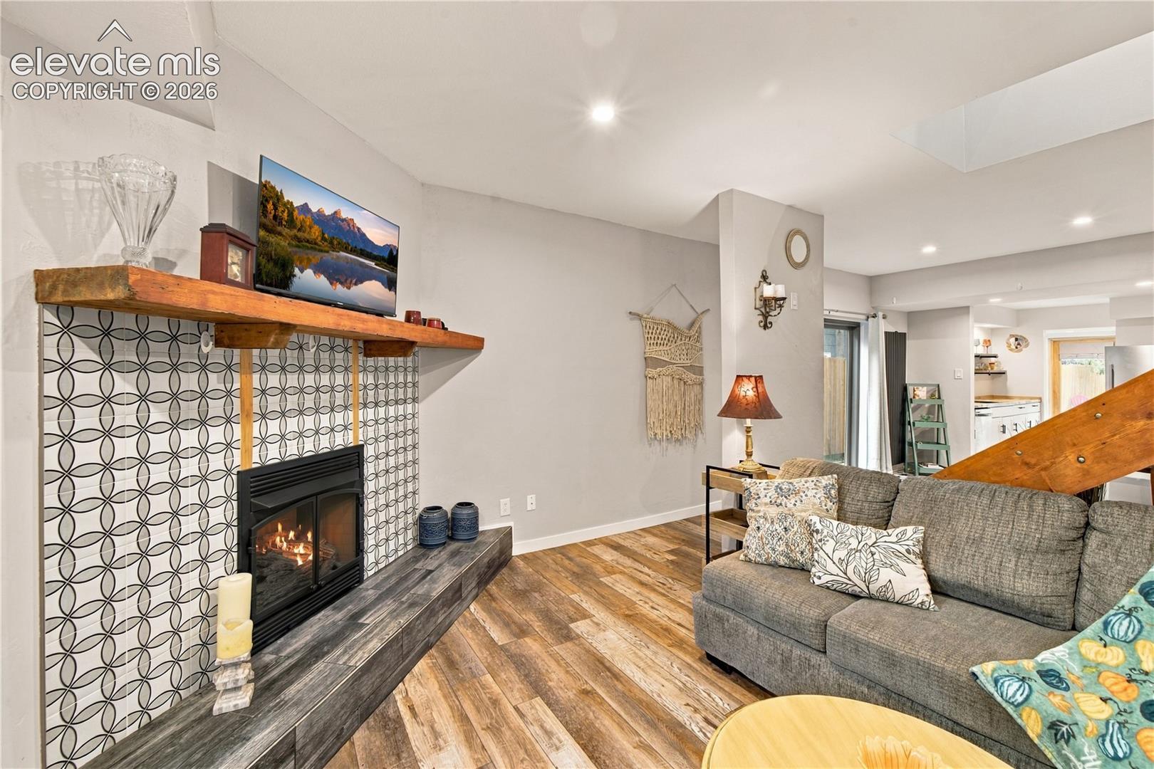 Fireplace featuring a patterned tile surround, a live-edge wood mantel, a black metal firebox, and a raised hearth finished in wood-finish planks, complemented by wood-finish flooring and recessed lighting
