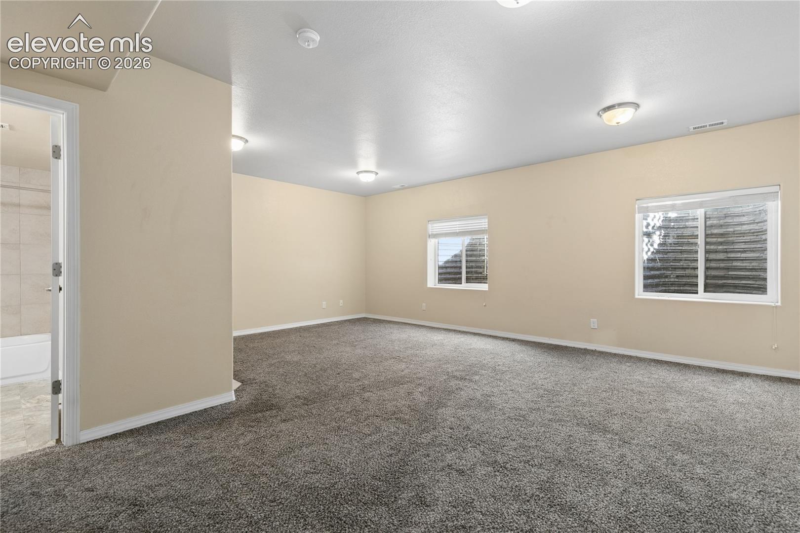 Expansive room featuring plush gray carpeting, two windows with white blinds, and recessed lighting fixtures