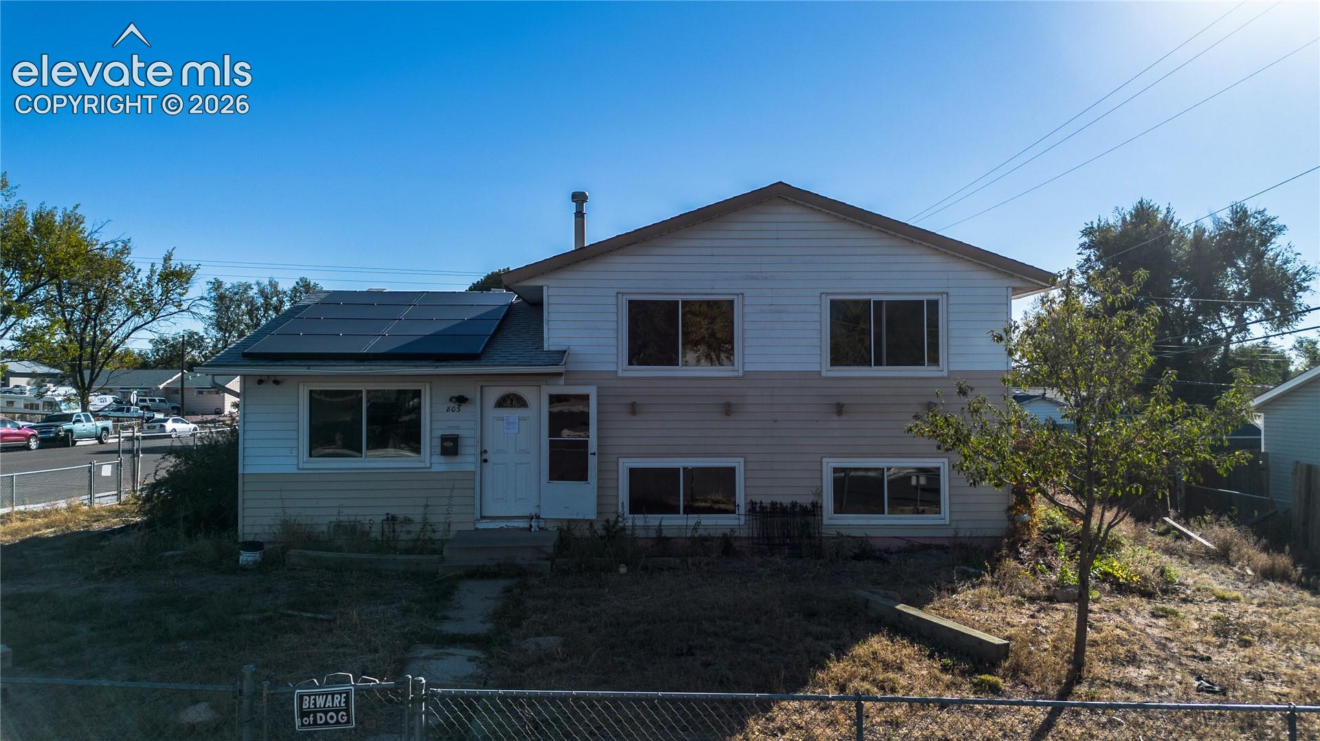 Two-story residence featuring horizontal siding, roof-mounted solar panels, multiple windows, and a chain-link fence perimeter