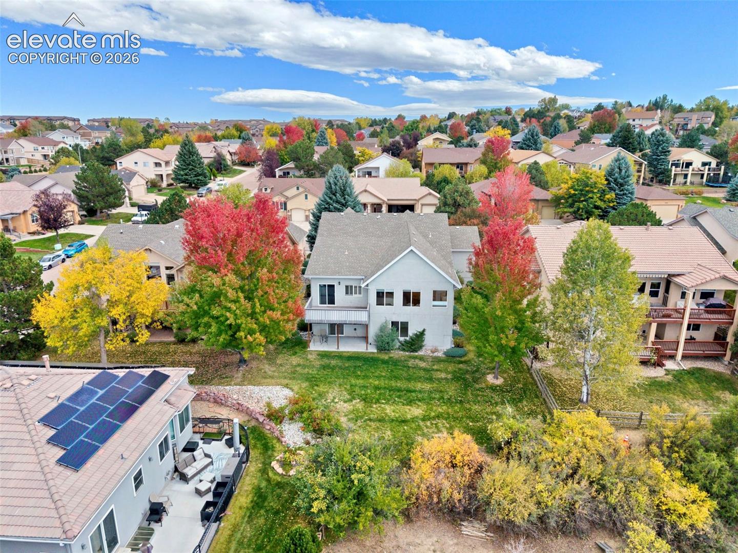 Aerial perspective featuring a multi-story residence with a pitched shingle roof, integrated solar panel array, and a rear concrete patio