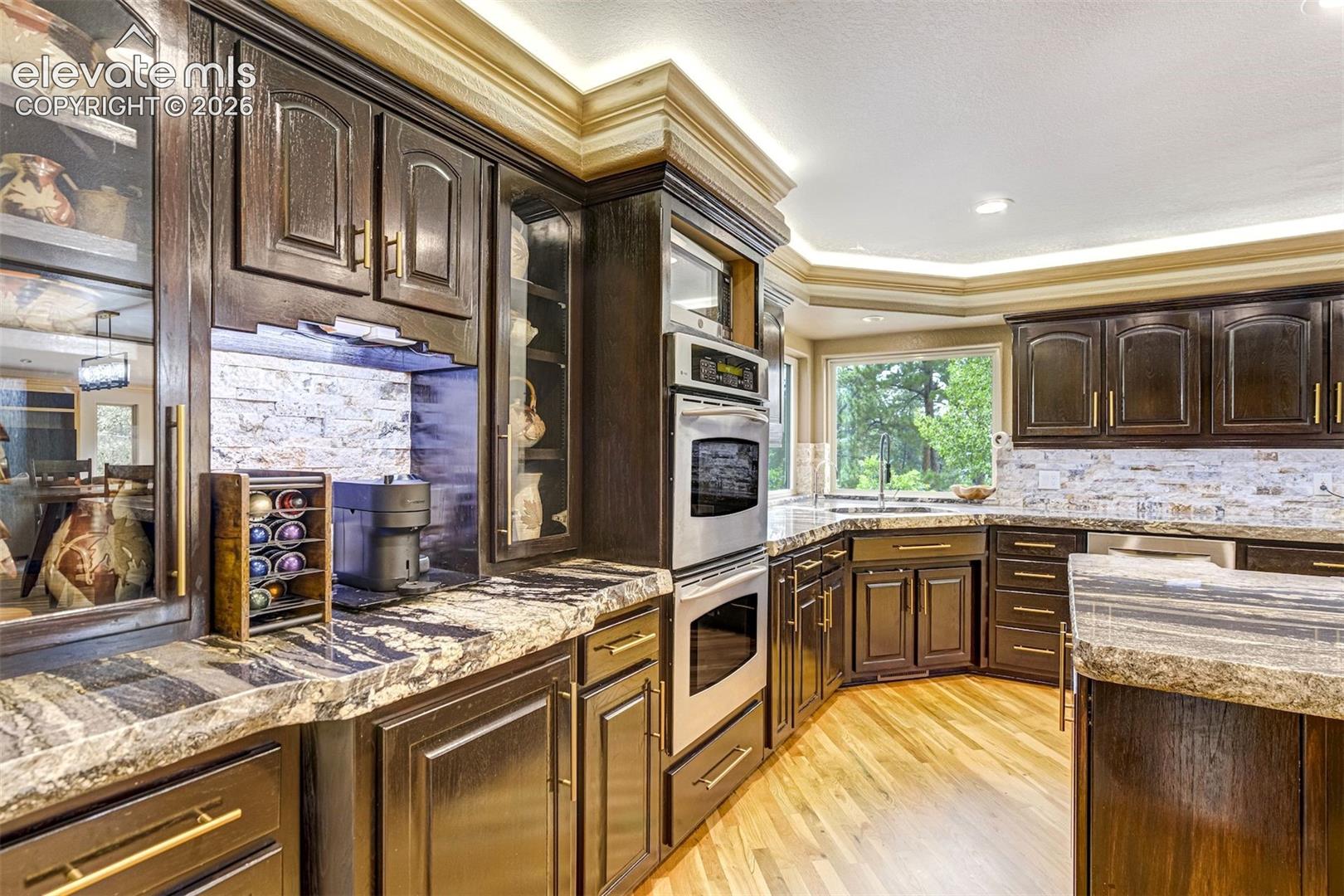 Dark wood cabinetry, stone countertops, stainless steel double oven, stainless steel microwave, and wood-finish flooring