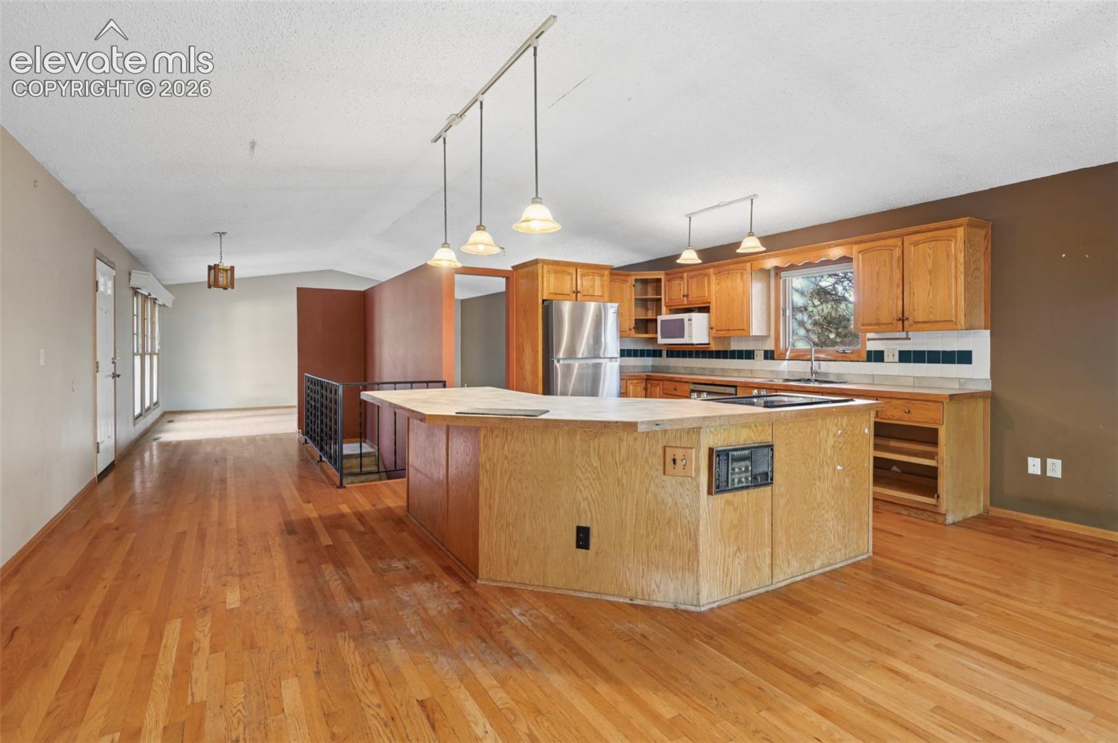 Hardwood flooring throughout, kitchen island with built-in cooktop, stainless steel refrigerator, wood cabinetry, and track lighting with pendant fixtures
