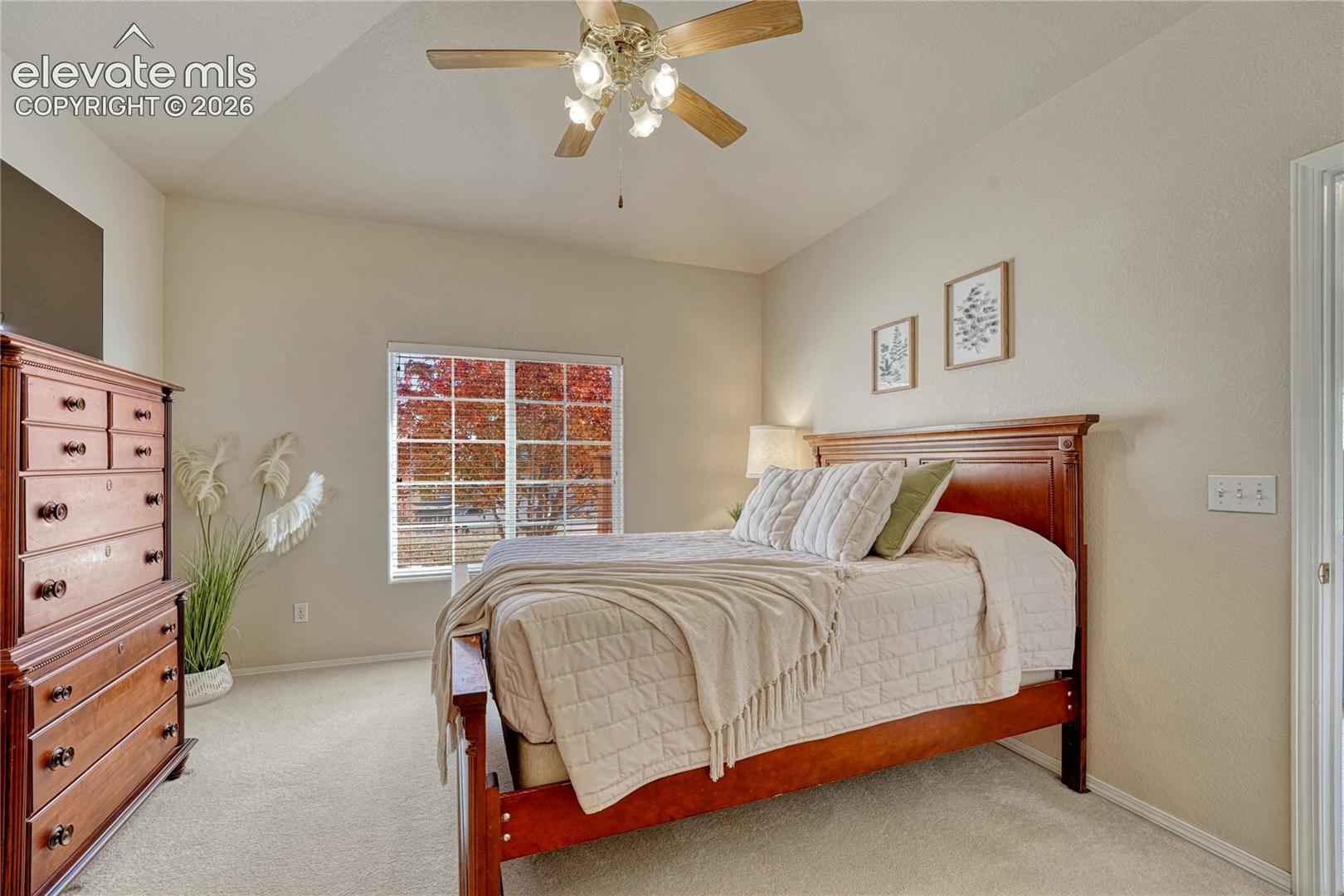 Bright natural light highlights the primary bedroom's vaulted design, ceiling fan, and serene setting overlooking mature trees
