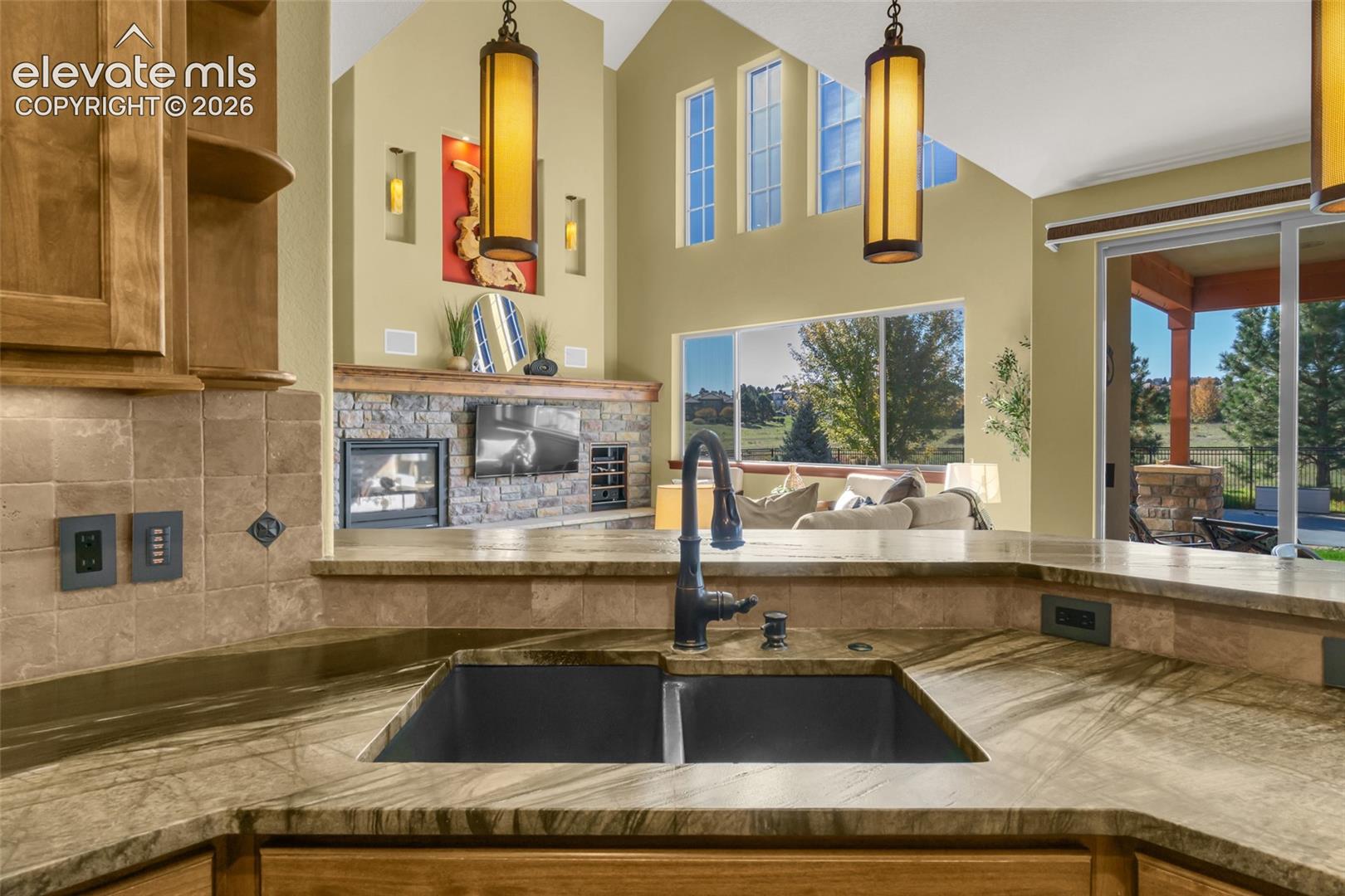 Kitchen featuring stone-surface countertops, dual basin undermount sink with a high-arch faucet, tile backsplash, vaulted ceiling, and stone-accented fireplace