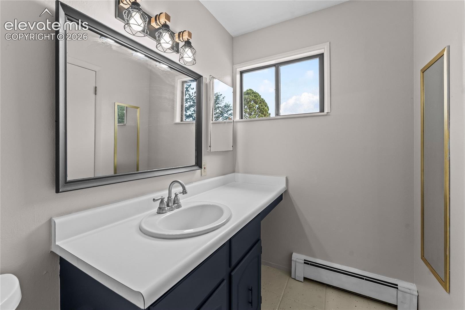 Vanity featuring a dark-toned cabinet, integrated basin, and wide countertop, illuminated by a multi-bulb industrial-style fixture above a framed mirror