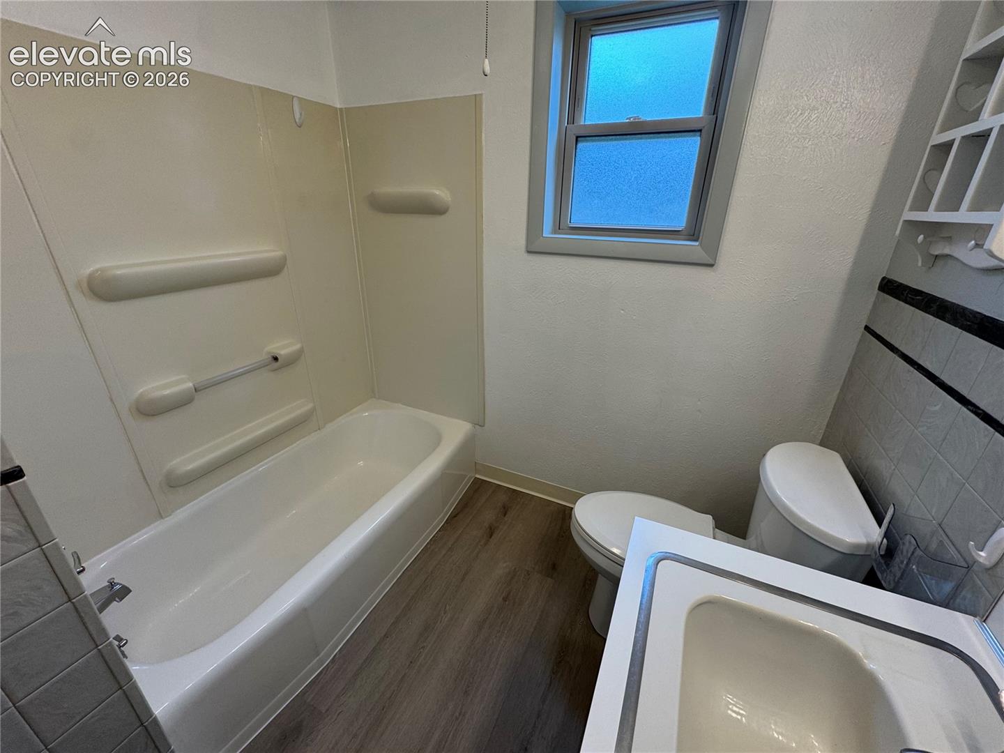 Bathroom featuring wood-finish flooring, a white bathtub with integrated surround, a single-basin vanity, and a window