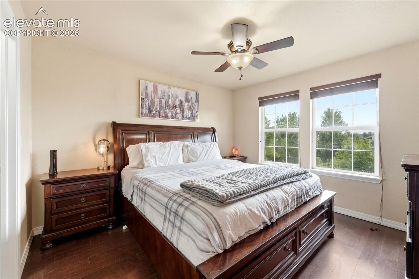 Wood-finish flooring, ceiling fan with integrated lighting, two windows with roller shades, and light-toned wall paint
