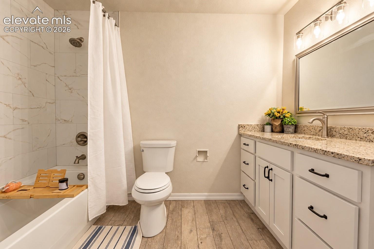 White vanity with black pulls, granite countertop, brushed nickel faucet, large mirror with a four-bulb light fixture, wood-finish flooring, and a shower-tub combination featuring white tiled walls
