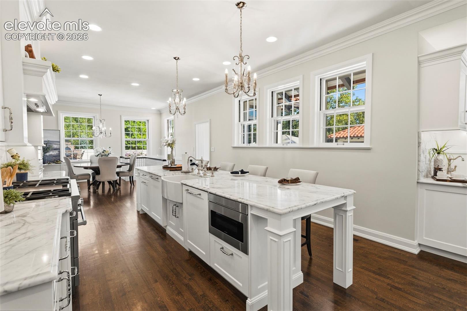 Kitchen featuring an expansive stone-topped island with an integrated microwave and farmhouse sink, wood-finish flooring, custom white cabinetry, and multi-light chandeliers