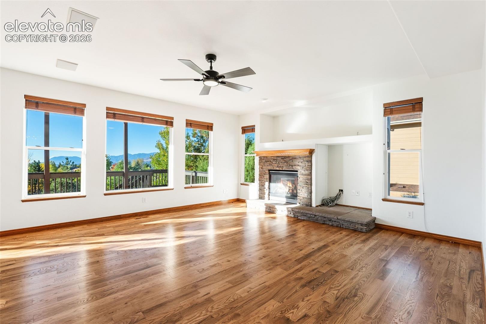 Open living space featuring wood-finish flooring, a fireplace with stone surround and wood mantel, multiple windows, a ceiling fan, and mountain scenery
