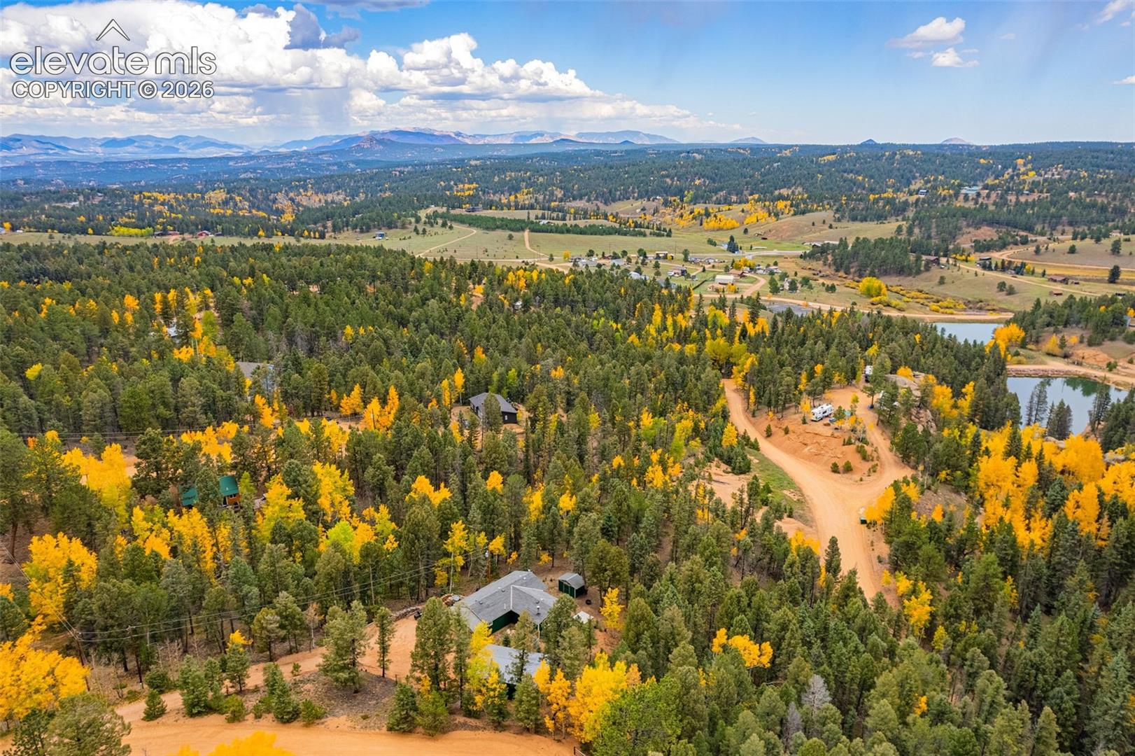 Aerial perspective of a multi-structure property nestled within a densely wooded landscape featuring mature evergreen and deciduous trees, expansive acreage, and mountainous terrain