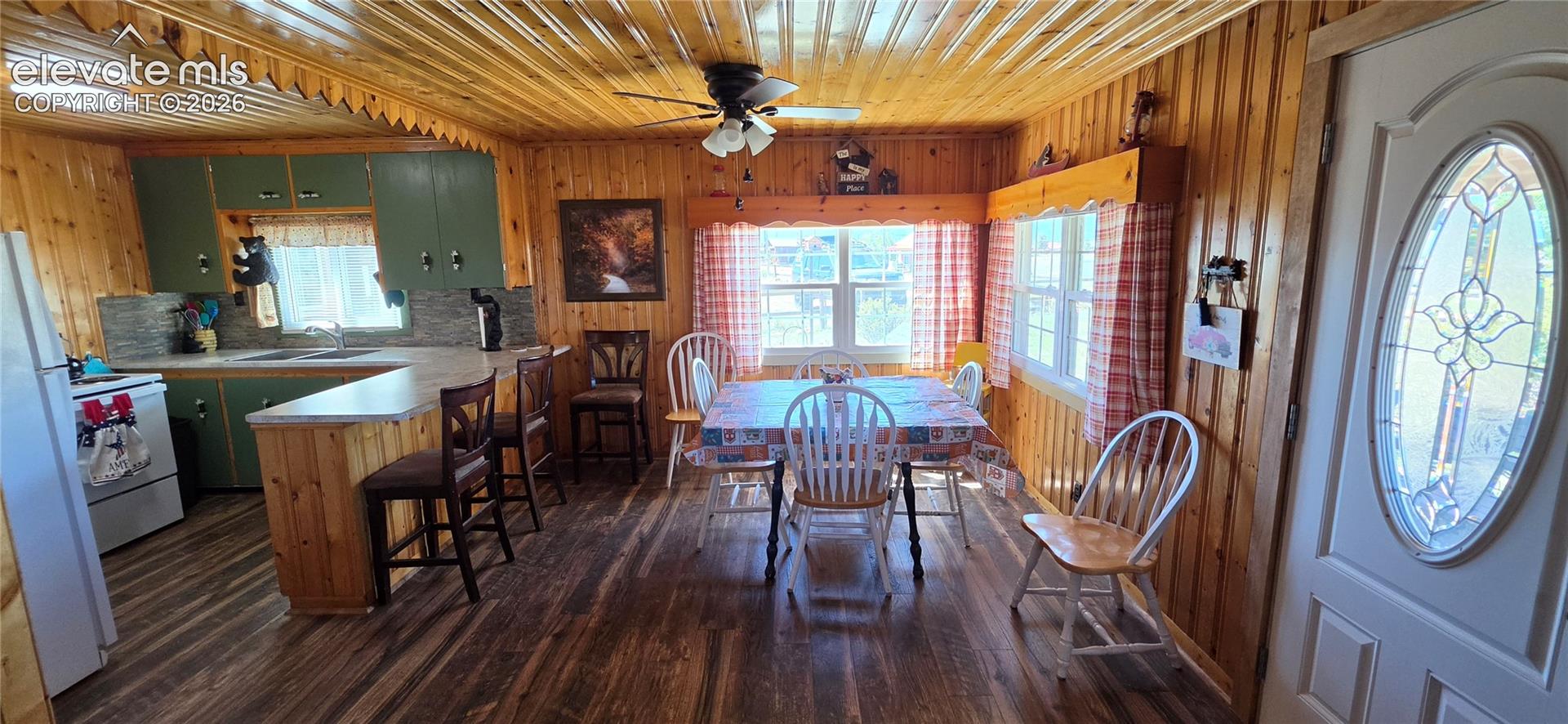 Wood-finish flooring throughout, wood-paneled walls and ceiling, kitchen with green cabinetry, breakfast bar, and decorative front door with oval stained glass