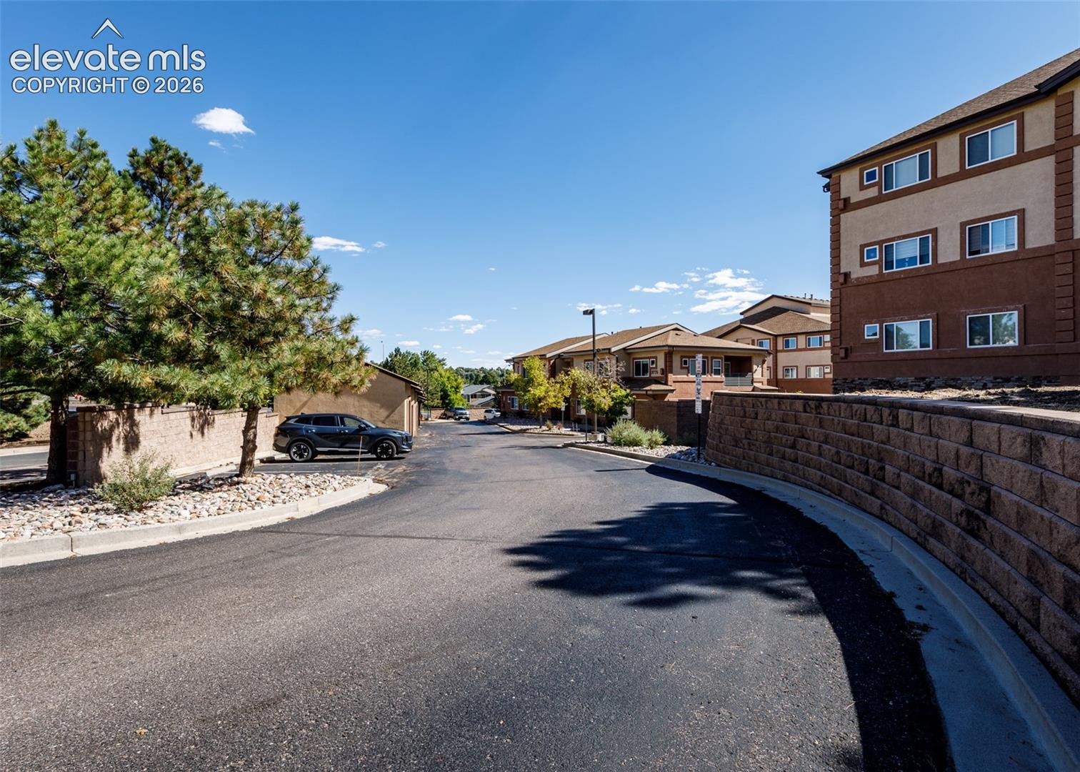 Exterior view featuring multi-story structures with earth-toned stucco siding, retaining walls constructed of stacked concrete masonry blocks, paved asphalt surfaces, and mature pine trees