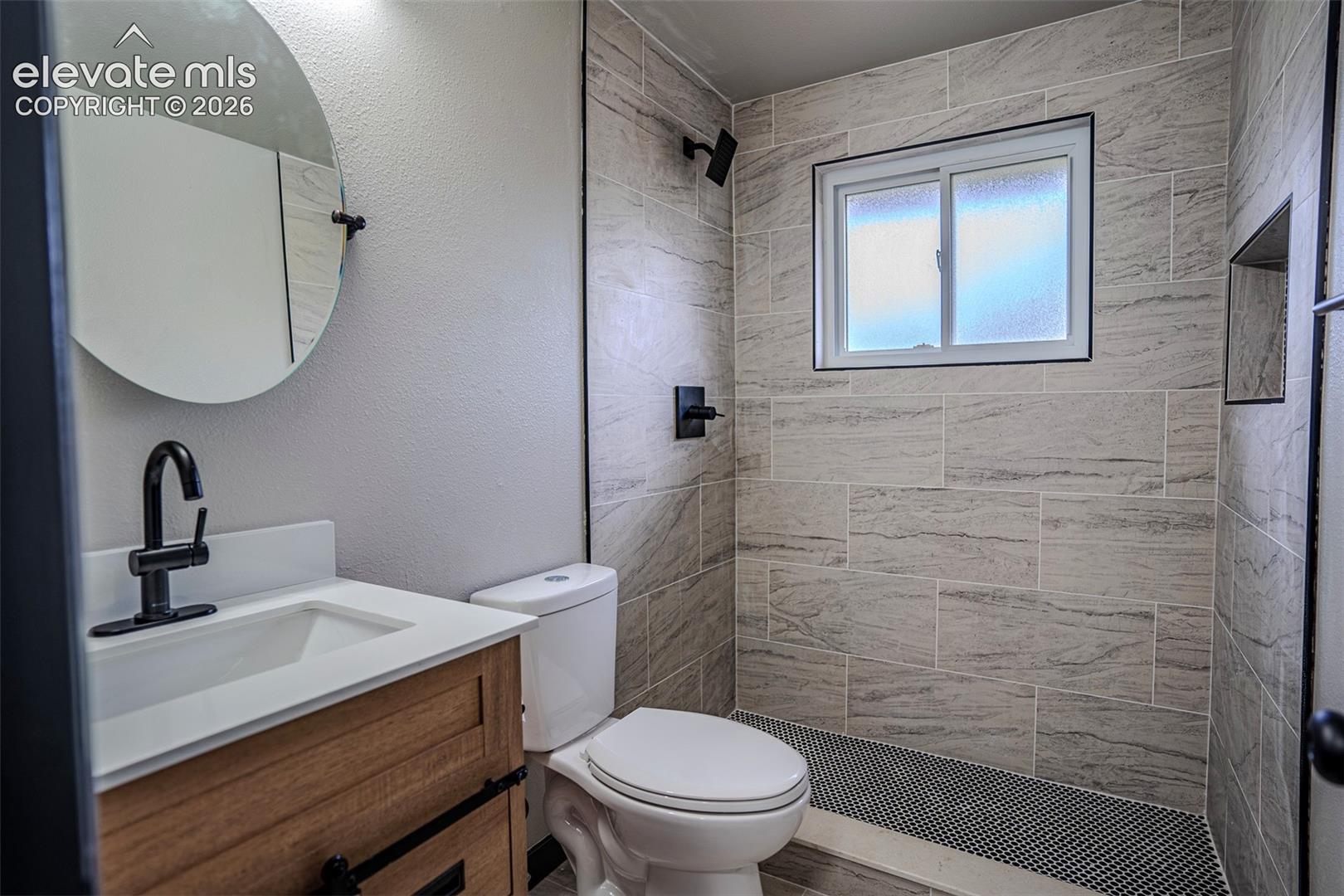 Bathroom featuring a wood-finish vanity with a white countertop, circular wall-mounted mirror, matte black plumbing fixtures, walk-in shower with tile surround, and a built-in recessed niche
