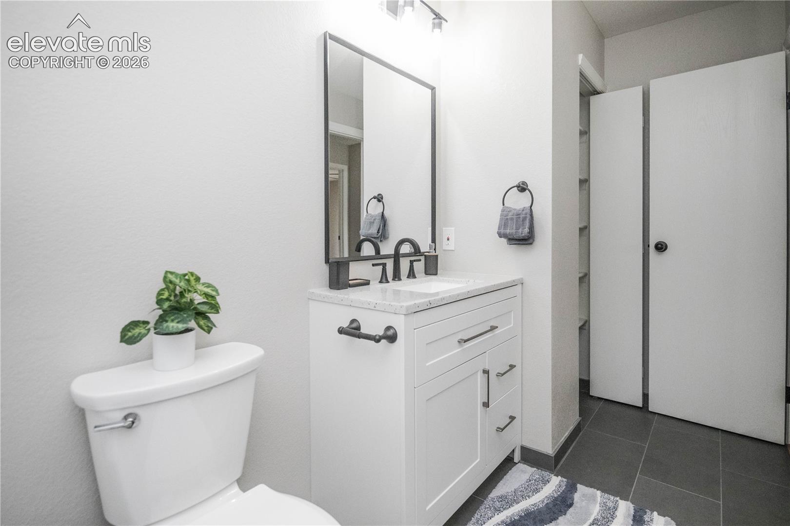 White vanity featuring a light-toned stone-finish countertop, rectangular under-mount sink, and matte black hardware, accented by a matching framed mirror and slate-tone tile flooring