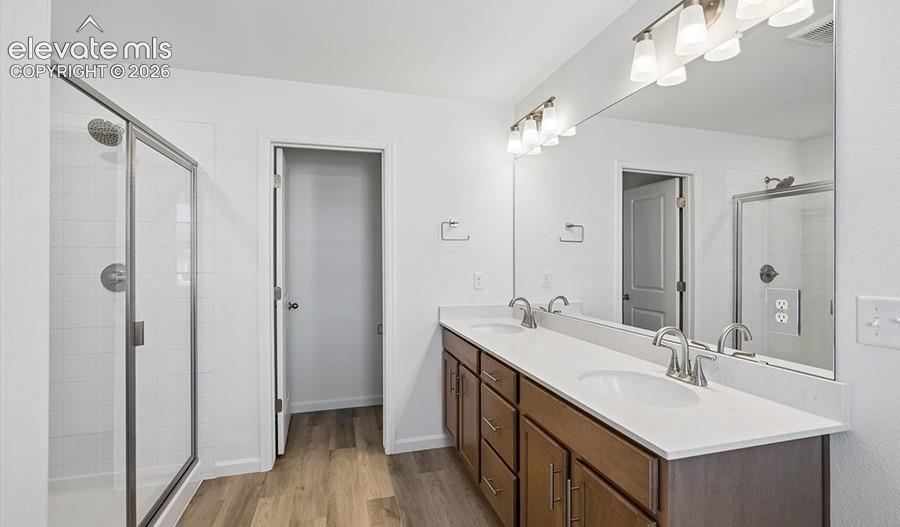 Double vanity featuring stone countertops, integrated dual sinks, wood-finish cabinetry, and a full-width vanity mirror, complemented by a walk-in shower with a glass enclosure and wood-finish flooring