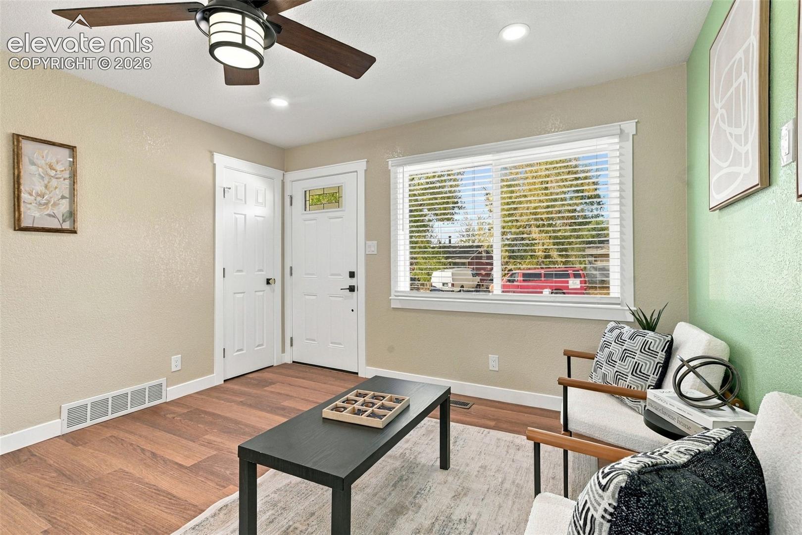 Interior space featuring wood-finish flooring, a multi-pane window with horizontal blinds, a ceiling fan with integrated lighting, and a six-panel entry door with a decorative glass transom