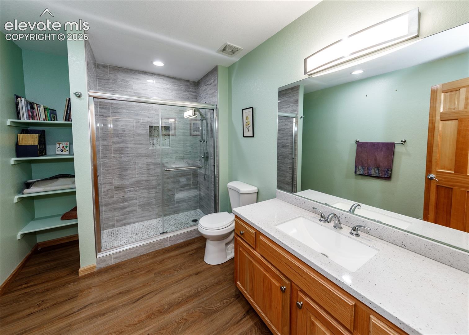 Wood-finish flooring, built-in shelving, glass-enclosed shower with tiled walls, vanity with a rectangular undermount sink, and a stone-finish countertop