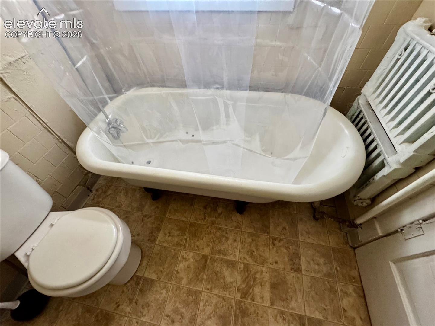 Clawfoot bathtub with chrome fixtures, white toilet, tiled flooring, and a window