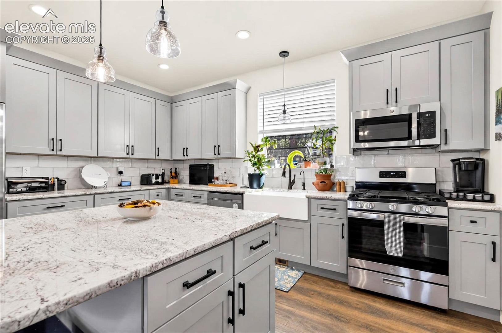 Kitchen featuring grey shaker-style cabinetry, a stone-finish island countertop, farmhouse-style sink, stainless steel appliances, and pendant lighting