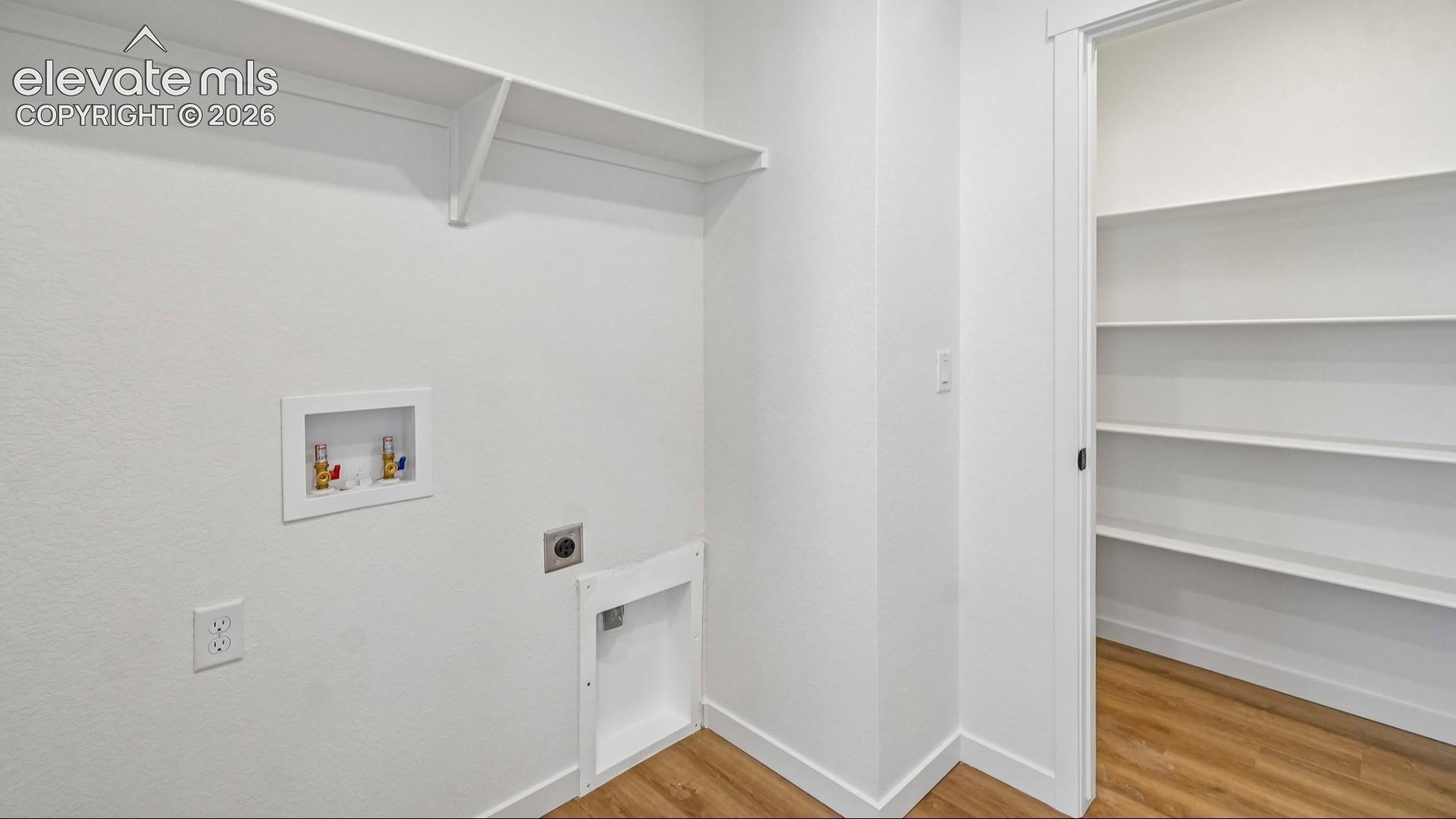 Utility area featuring wood-finish flooring, a wall-mounted shelf, and recessed water and dryer hookups, including a pantry area with integrated tiered shelving