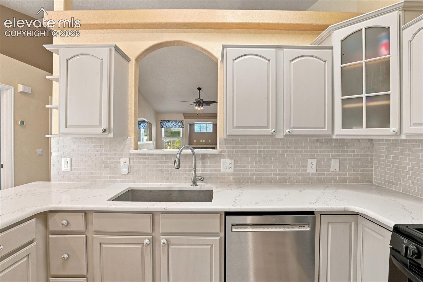 Kitchen featuring light-toned cabinetry, stone-finish countertops, subway tile backsplash, arched pass-through window, and stainless steel undermount sink with a gooseneck faucet