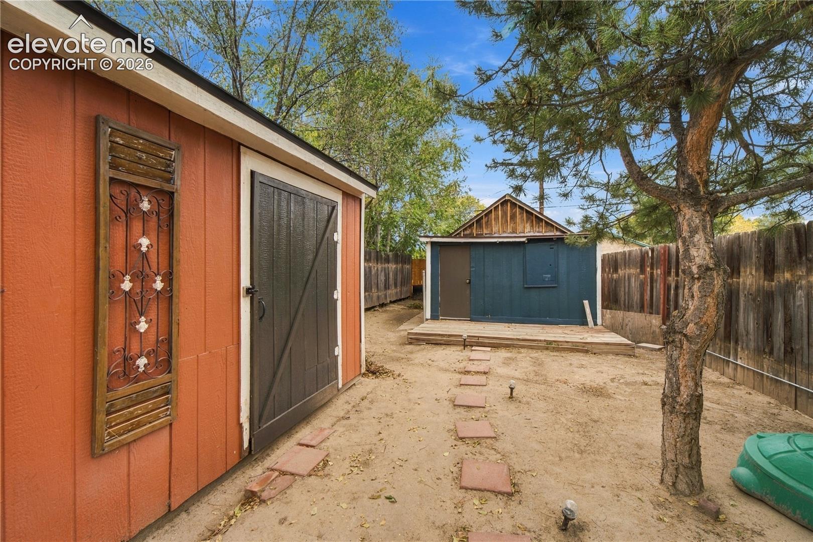 Exterior area featuring two detached outbuildings, wood-style fencing, a stone paver path, and mature trees