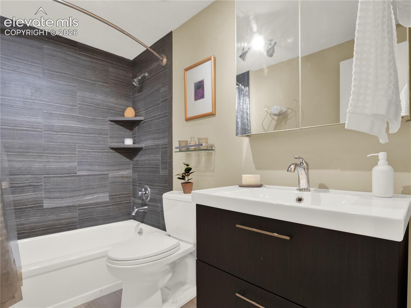 Bathroom featuring a dark wood-finish vanity with a white rectangular sink, a mirrored medicine cabinet, a tub-shower combination with floor-to-ceiling charcoal tile surround, and integrated wall shelving
