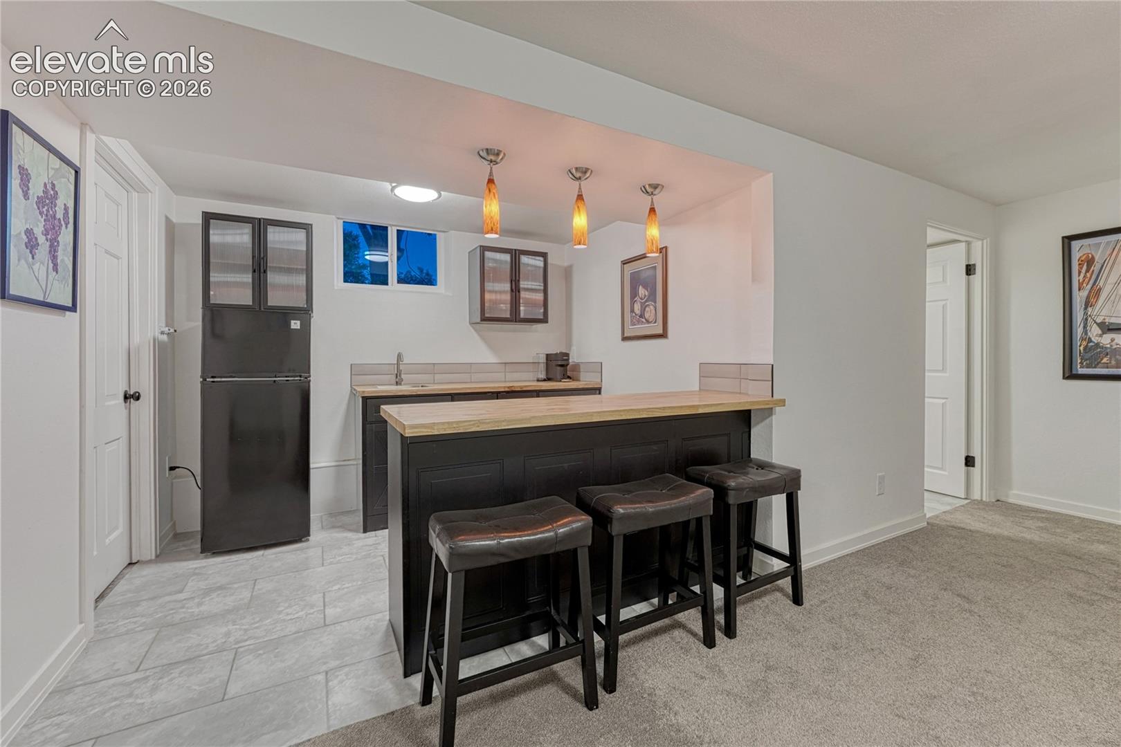 Wet bar featuring custom dark cabinetry, wood-finish countertop, tiled backsplash, integrated sink, and three pendant light fixtures