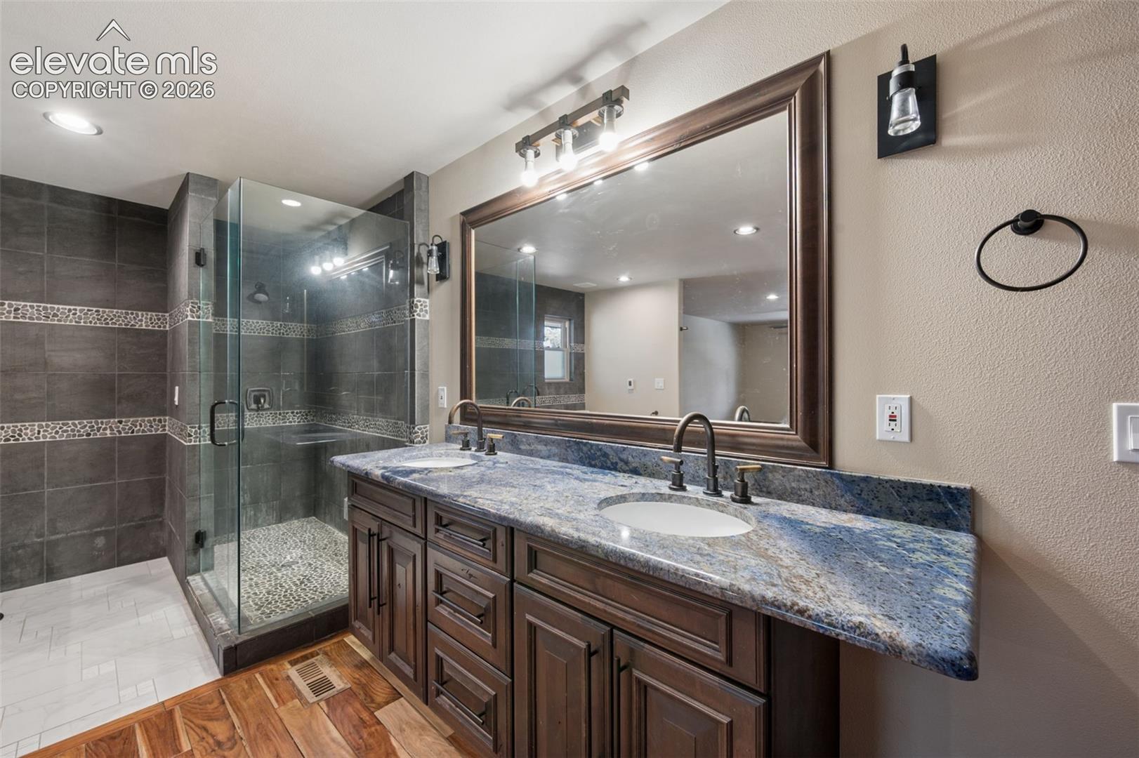 Double vanity featuring a blue-toned stone countertop, dual undermount sinks, custom dark wood cabinetry, and a glass-enclosed shower with tiled walls and decorative stone accents