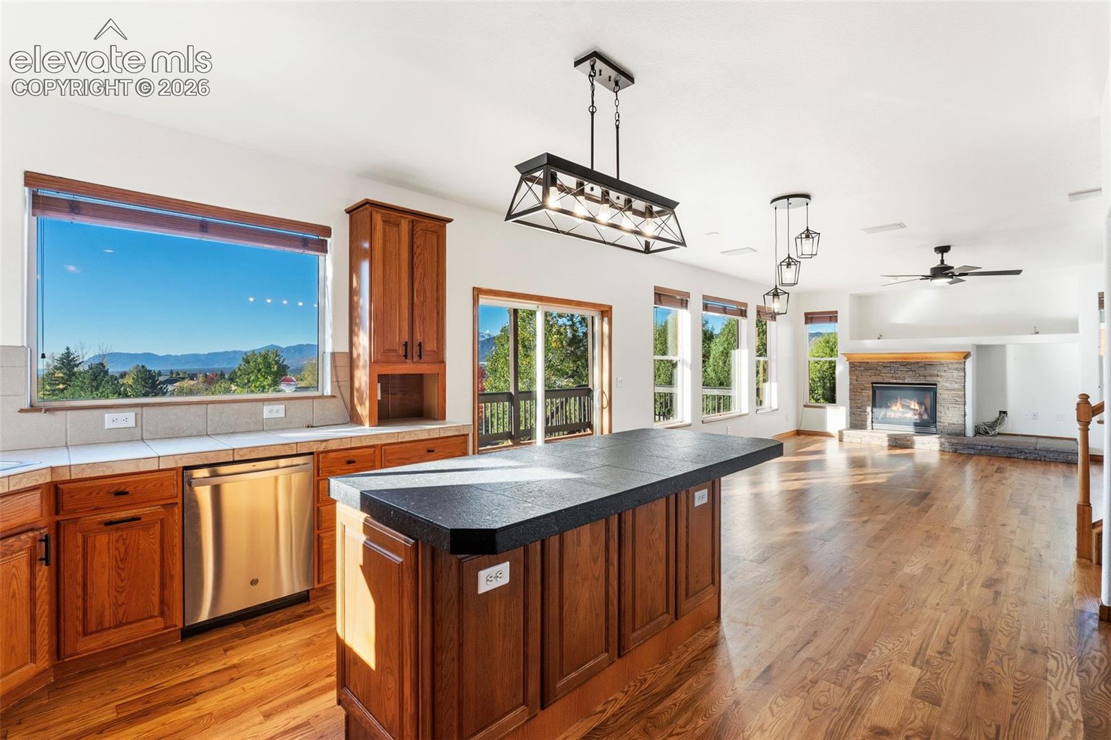 Kitchen featuring wood cabinetry, dark tile countertops, wood-finish flooring, a rectangular pendant light fixture, and a stone-surround fireplace