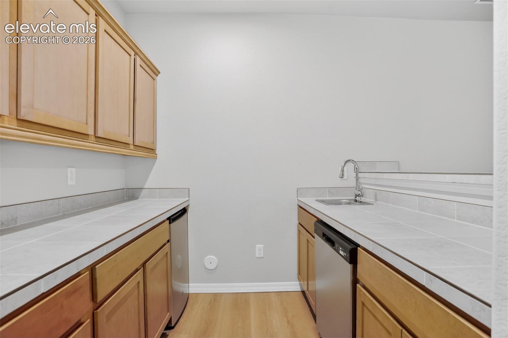 Galley-style kitchenette featuring wood-finish cabinetry, tile countertops with a matching backsplash, stainless steel sink with a chrome faucet, and integrated stainless steel appliances