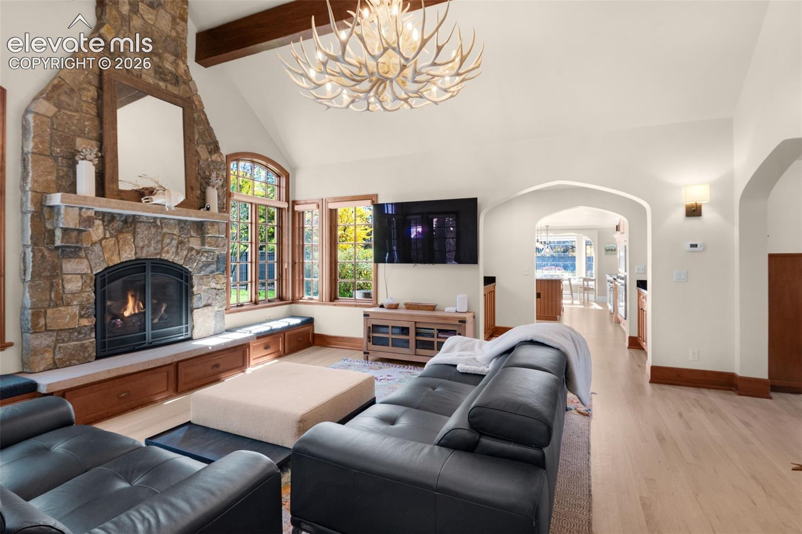 Vaulted ceiling with exposed wood beam, floor-to-ceiling stone fireplace, built-in wooden cabinetry, arched architectural openings, and light-tone wood-finish flooring