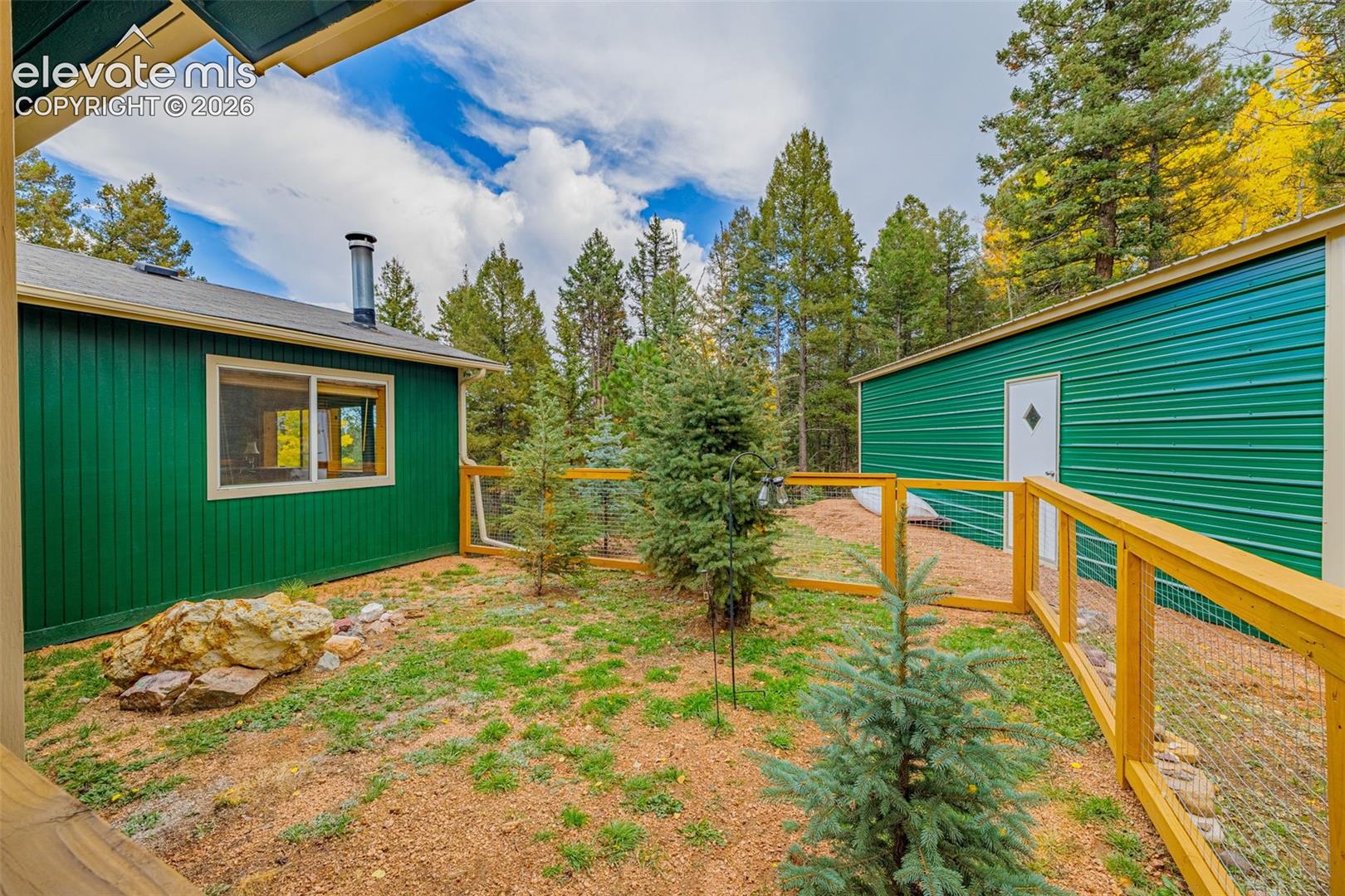Exterior courtyard featuring green vertical siding, a metal-sided detached structure, wood-framed wire fencing, and mature evergreen landscaping