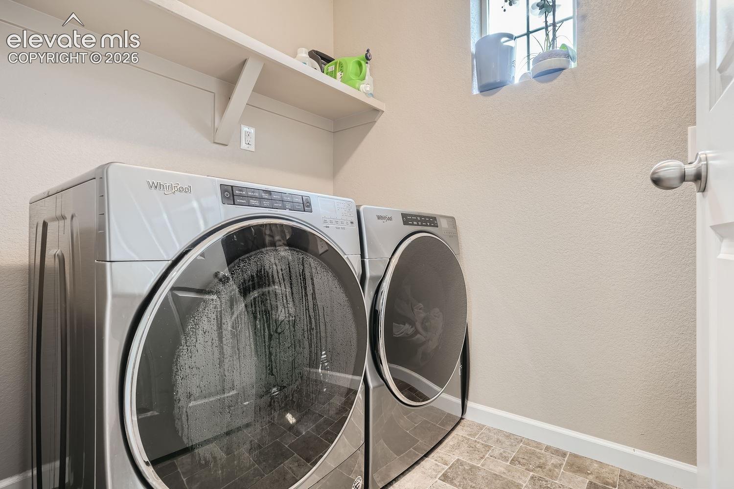 Laundry room featuring stainless steel front-load appliances, a built-in overhead shelf, a high-set window, and stone-tile flooring