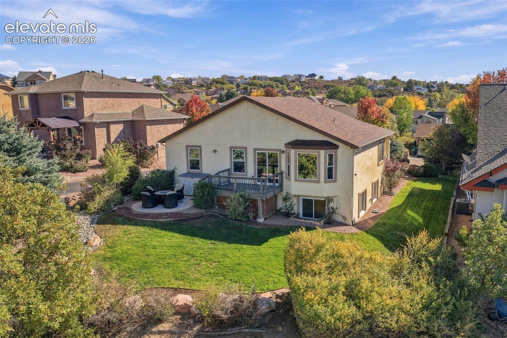 Stucco exterior featuring a raised wooden deck, ground-level stone patio, manicured lawn, and mature landscaping