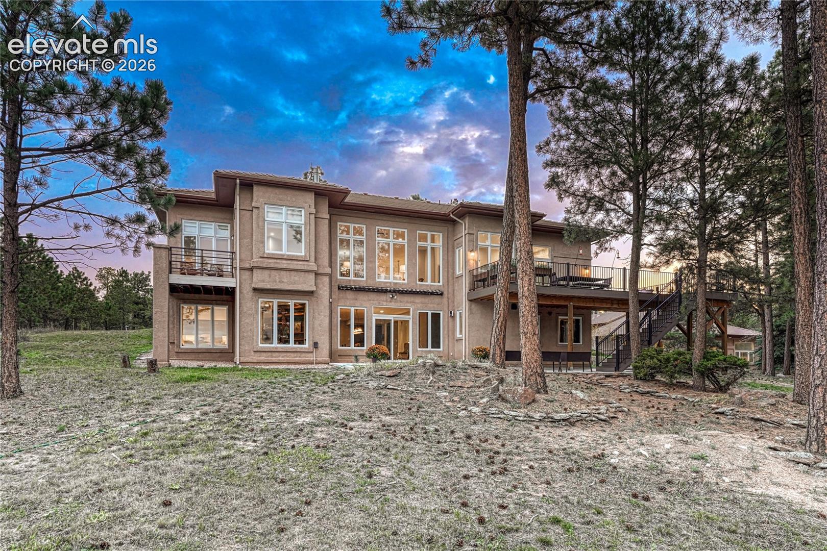 Multi-story stucco exterior featuring an elevated deck with metal railings, an upper-level balcony, expansive multi-pane windows, and a mature treed landscape