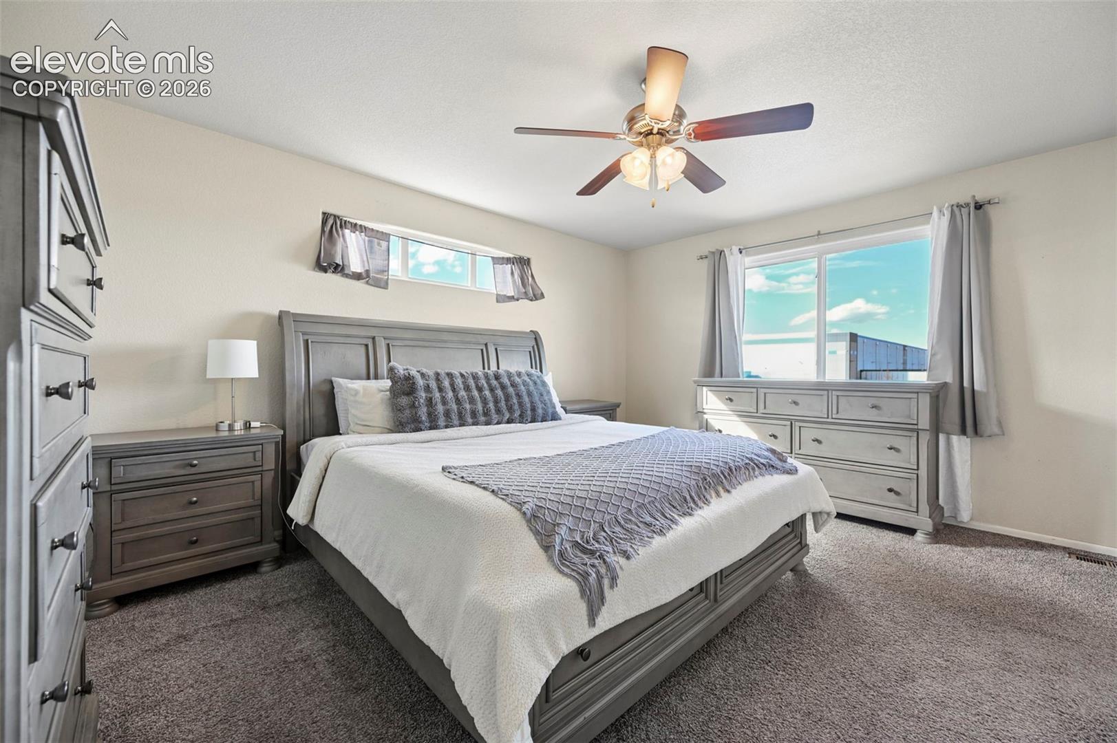 Bedroom featuring neutral-toned carpeting, a multi-blade ceiling fan with integrated lighting, a sliding window, and an elongated transom window