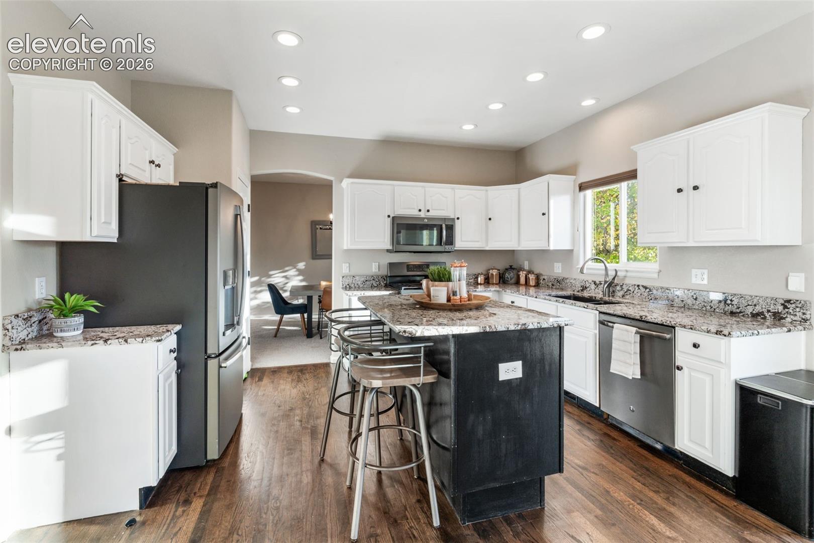 Kitchen featuring white shaker-style cabinetry, stone-surface countertops, a contrasting central island, stainless steel appliances, and wood-finish flooring